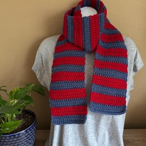 May include: A hand-crocheted scarf featuring horizontal stripes in alternating vibrant red and dark gray yarn. The scarf is draped around the neck of a display form wearing a plain light gray t-shirt, showcasing the textured stitch pattern.
