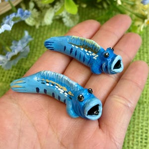 May include: Two hand-painted, blue fish figurines with open mouths and black eyes. The fish have white and gold accents along their backs and fins. The figurines are held in a person's hand, with a background of flowers.