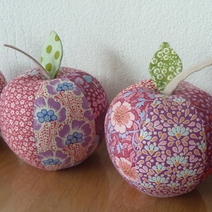 Könnte beinhalten: Vier Stoffäpfel mit Stielen und Blättern. Die Äpfel bestehen aus gemustertem Stoff in Rot-, Rosa- und Lilatönen. Der Stoff hat florale Designs. Die Äpfel sind auf einer Holzoberfläche angeordnet.