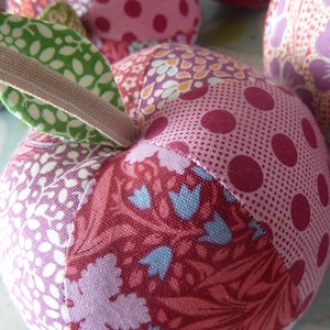 Könnte beinhalten: Nahaufnahme von mehreren Stoff-Apfel-Ornamente. Die Äpfel sind aus Patchwork-Stoff in Rosa-, Rot- und Lilatönen mit Blumen- und Tupfenmustern gefertigt. Jeder Apfel hat ein grünes Stoffblatt und einen beigen Stiel.