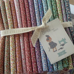 Könnte beinhalten: Ein Bündel Stoffstreifen mit Blumenmuster in verschiedenen Farben, gebunden mit einem Band mit der Aufschrift "Quilt Collection". Ein Etikett zeigt "Brie... Basics" mit einer Cartoon-Illustration eines Mädchens und einer Nähmaschine. Stoffmuster und eine Karten mit Knöpfen sind sichtbar.