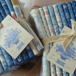 Könnte beinhalten: Mehrere Bündel blau-weiß gemusterter Stoffe, gebunden mit cremefarbenem Band. Jedes Bündel hat ein Etikett mit "SOMETHING BLUE" und einem Blumendesign, der Marke "Tilda". Die Stoffmuster umfassen Blumen, Punkte und Streifen.