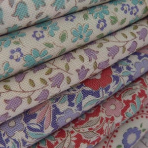 Könnte beinhalten: Ein Stapel gefalteter Stoffstücke mit Blumenmustern in Blau-, Lila-, Rot- und Grüntönen auf cremefarbenem Grund. Ein beiges Stoffband mit dem sichtbaren Text "QUILT COLLECTION" umschließt den Stapel der gefalteten Textilien.
