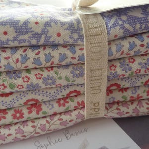 Könnte beinhalten: Ein Stapel aus fünf gefalteten Stoffstücken mit verschiedenen Blumenmustern in Blau-, Rot- und Rosatönen auf cremefarbenem Grund. Ein beiges Stoffband mit der Aufschrift Tilda Quilt Collection umschließt die Mitte des Stoffstapels.
