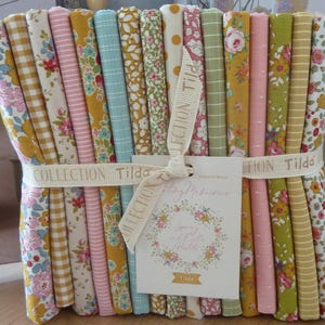 Könnte beinhalten: Ein Bündel bunter Stoffstreifen mit verschiedenen Blumen- und Mustermotiven, gebunden mit einem Band mit der Aufschrift "Collection Tilda". Ein Etikett mit floralem Kranz und Text "Spring and Easter Fabrics" ist angebracht.