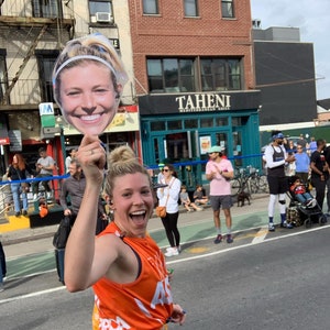 Puede incluir: Una mujer con una camiseta de running naranja sostiene un recorte de cart&oacute;n de la cara de una mujer. El recorte sonr&iacute;e y lleva una diadema. La mujer que sostiene el recorte sonr&iacute;e y lleva una diadema negra. El fondo es una calle de la ciudad con edificios y gente.