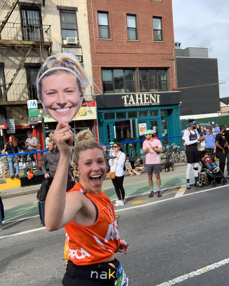 Puede incluir: Una mujer con una camiseta de running naranja sostiene un recorte de cart&oacute;n de la cara de una mujer. El recorte sonr&iacute;e y lleva una diadema. La mujer que sostiene el recorte sonr&iacute;e y lleva una diadema negra. El fondo es una calle de la ciudad con edificios y gente.
