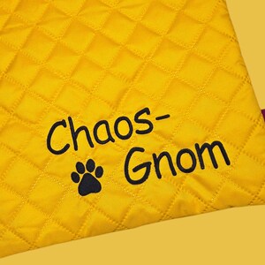 May include: A yellow quilted pet bed with the words "Chaos-Gnom" and a paw print in black. The bed has a diamond pattern and a small purple tag with white text.