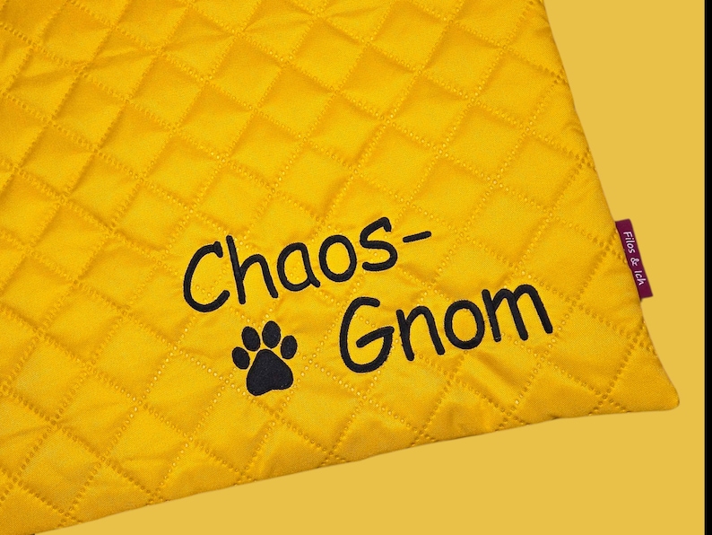 May include: A yellow quilted pet bed with the words "Chaos-Gnom" and a paw print in black. The bed has a diamond pattern and a small purple tag with white text.