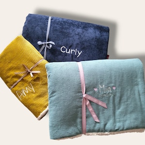May include: Three personalized corduroy blankets with white embroidery. The blankets are folded and tied with ribbon. The blankets are blue, green, and yellow. The names "Curly", "Amy", and "Mia" are embroidered on the blankets.