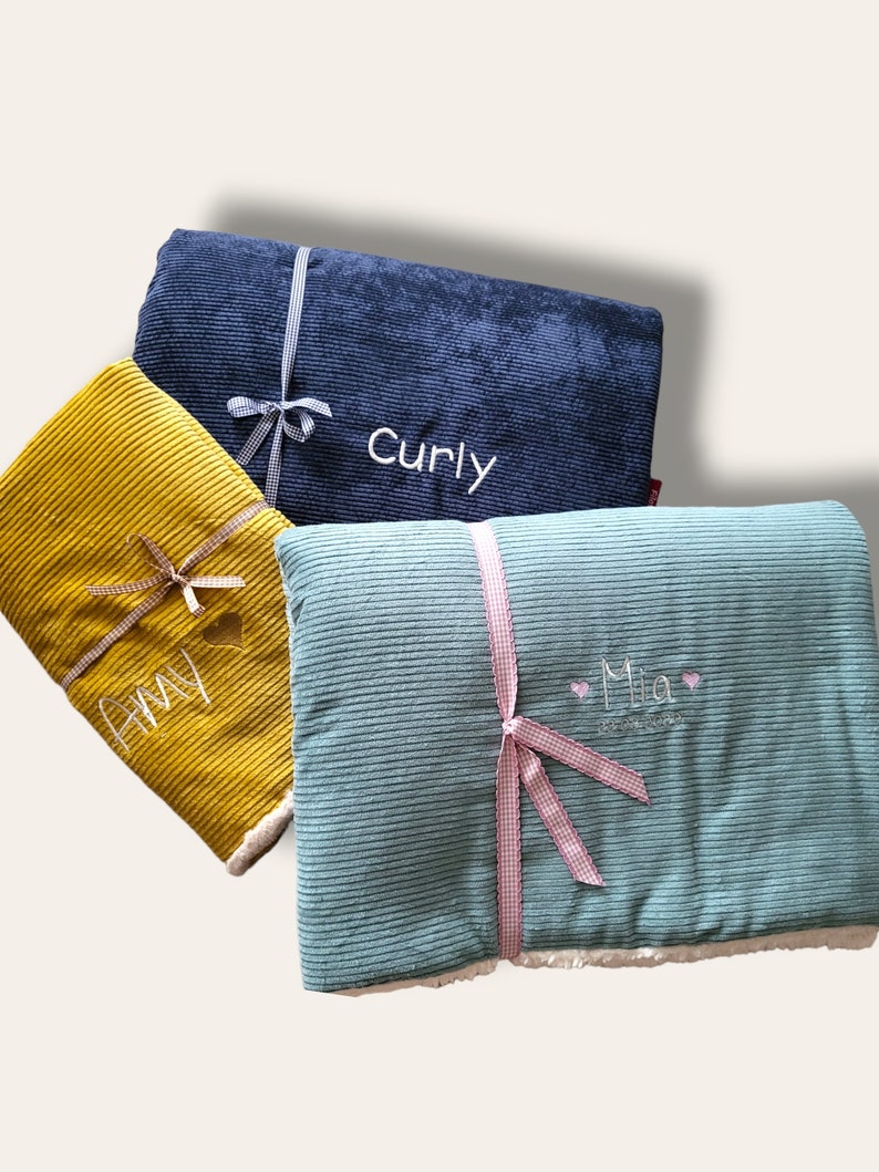 May include: Three personalized corduroy blankets with white embroidery. The blankets are folded and tied with ribbon. The blankets are blue, green, and yellow. The names "Curly", "Amy", and "Mia" are embroidered on the blankets.