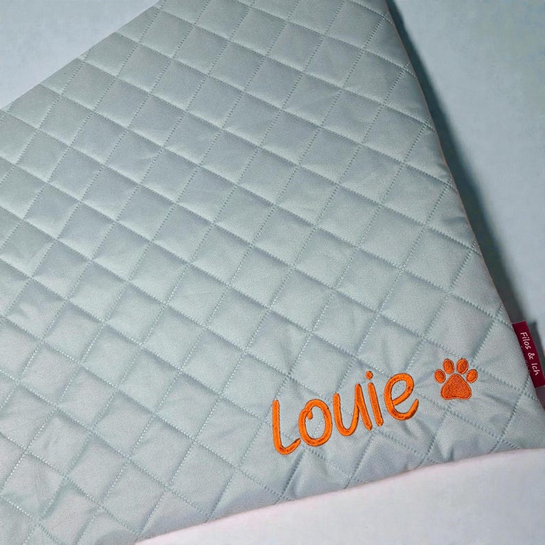 May include: A light blue, quilted pet bandana with orange embroidered text that reads "Louie" and a paw print. The bandana has a diamond pattern and a red tag that reads "Filos & Ich".