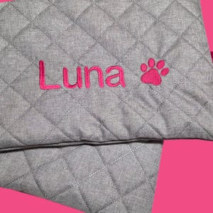 May include: Gray quilted pet bed with the name "Luna" embroidered in pink, along with a pink paw print. A pink tag with the text "Filos & Ich" is attached to the side. The bed has a diamond pattern.