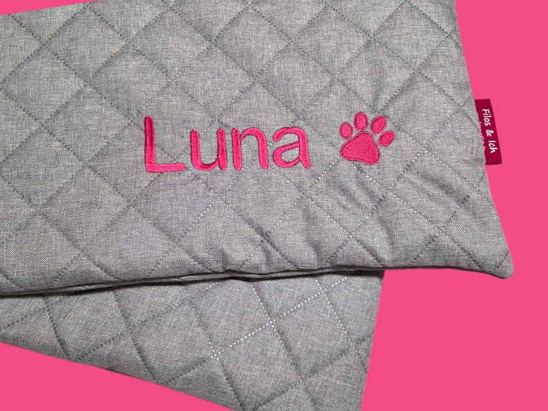 May include: Gray quilted pet bed with the name "Luna" embroidered in pink, along with a pink paw print. A pink tag with the text "Filos & Ich" is attached to the side. The bed has a diamond pattern.