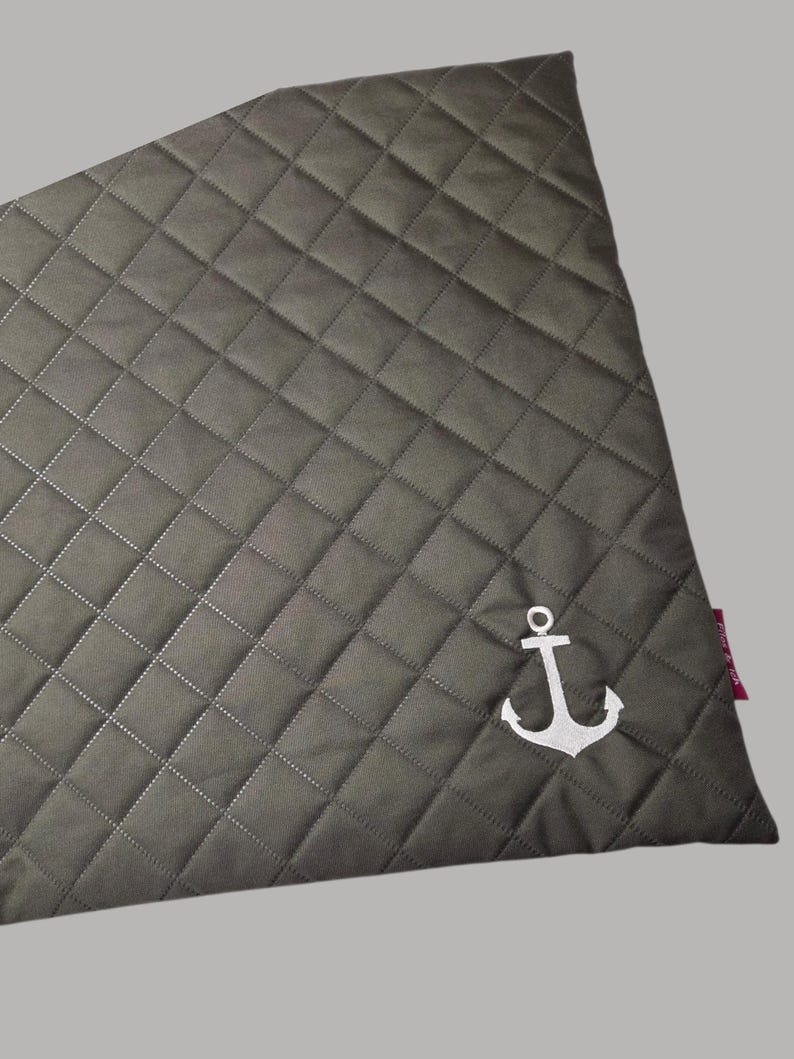 May include: A gray quilted fabric with a diamond pattern and white stitching. A white anchor emblem is embroidered in the lower right corner. The fabric has a tag on the right side.