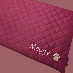 May include: A burgundy quilted pet bed with the name "Maggy" embroidered in white script. A tan paw print is embroidered near the bottom right corner. The bed has a diamond pattern.