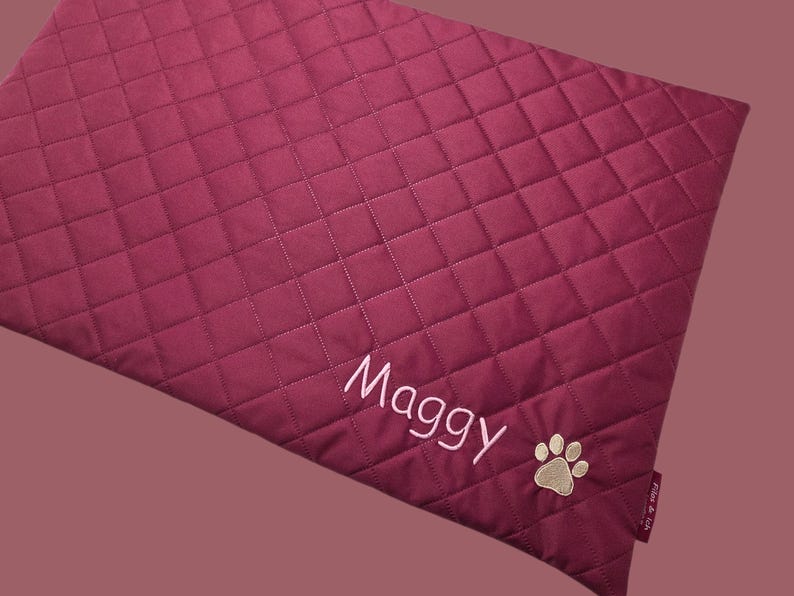 May include: A burgundy quilted pet bed with the name "Maggy" embroidered in white script. A tan paw print is embroidered near the bottom right corner. The bed has a diamond pattern.