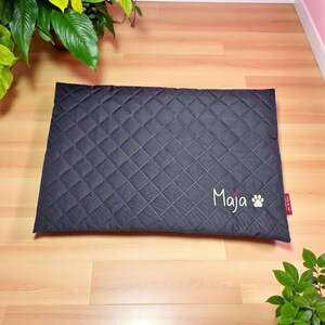 May include: A dark gray, quilted pet bed with a diamond pattern. The name "Maja" is embroidered in white script, along with a paw print. A small red tag is attached to the side. The bed is rectangular.