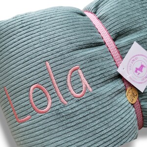 May include: A teal blue corduroy pillow with pink stitching and the name "Lola" embroidered in pink. The pillow has a pink and white gingham ribbon tied around it.