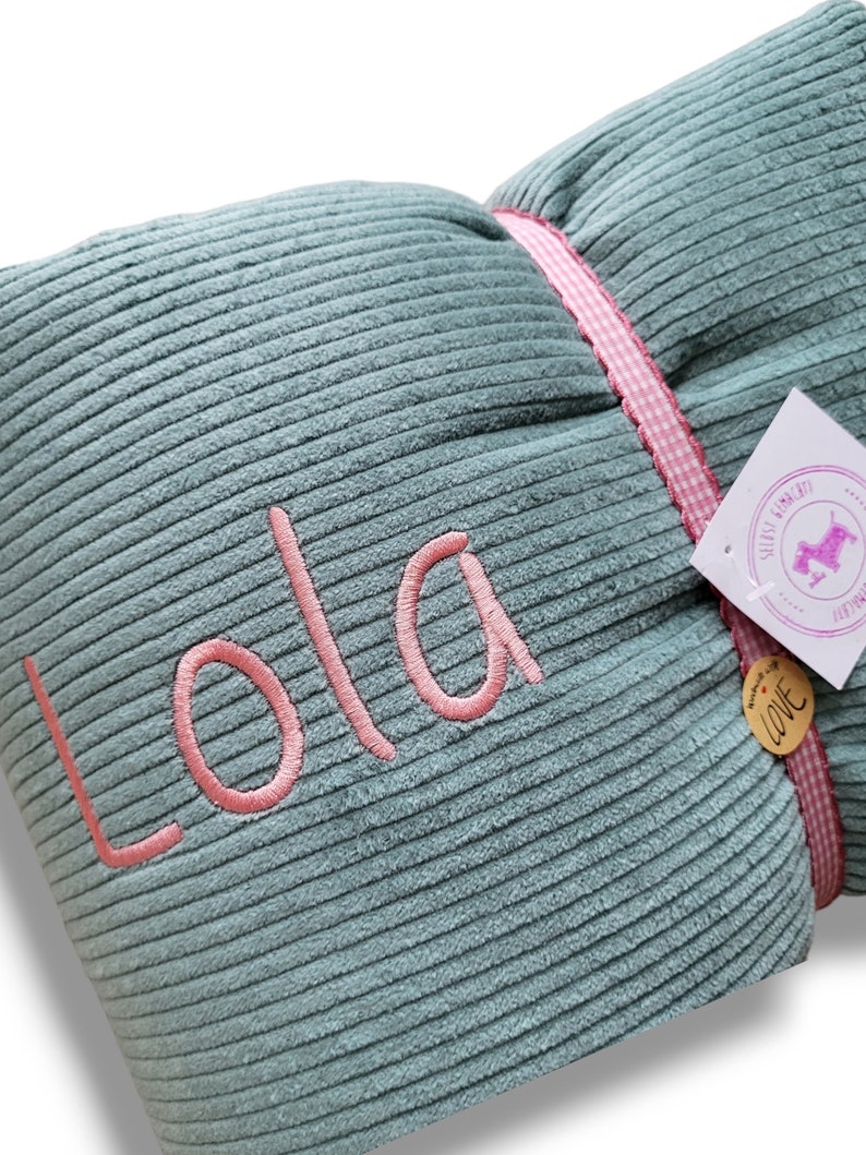 May include: A teal blue corduroy pillow with pink stitching and the name "Lola" embroidered in pink. The pillow has a pink and white gingham ribbon tied around it.