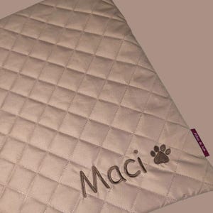 May include: A beige, quilted pet blanket with the name "Maci" embroidered in brown thread, along with a paw print. The blanket has a diamond pattern and a small purple tag that says "Files & leh".