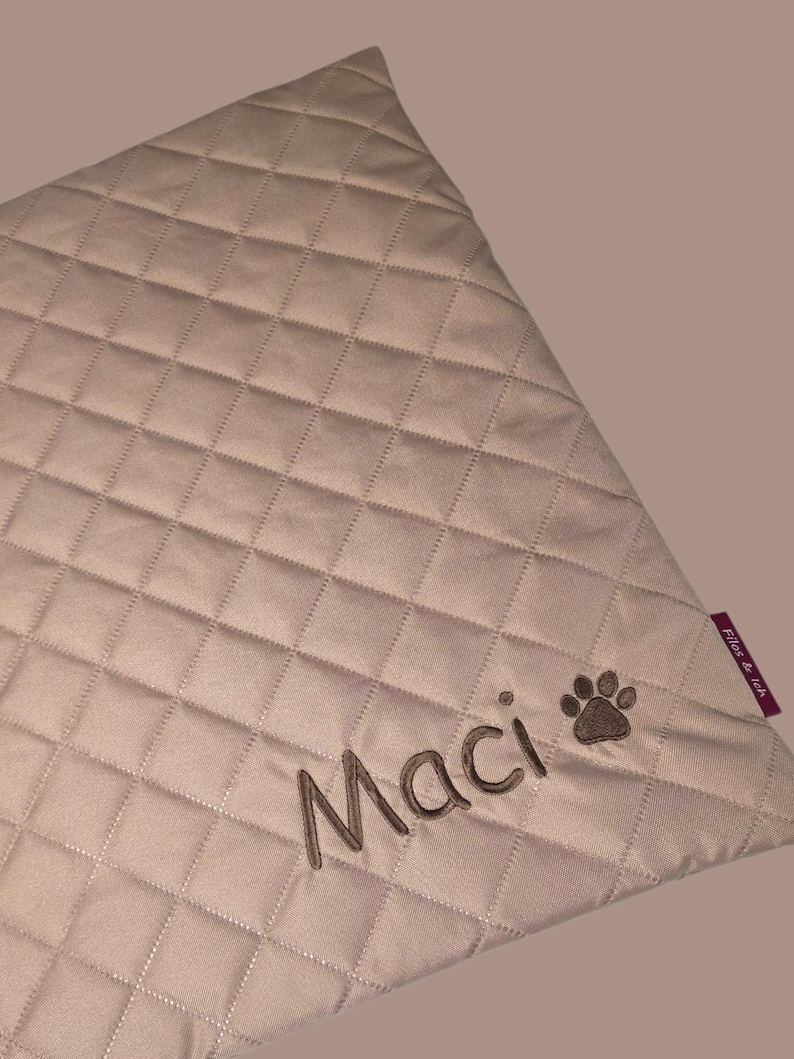 May include: A beige, quilted pet blanket with the name "Maci" embroidered in brown thread, along with a paw print. The blanket has a diamond pattern and a small purple tag that says "Files & leh".