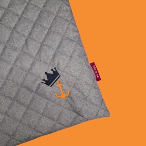May include: Gray quilted fabric with a navy blue crown and orange anchor embroidery. A red tag with the text "Fios & Ich" is attached to the corner. The background is orange.