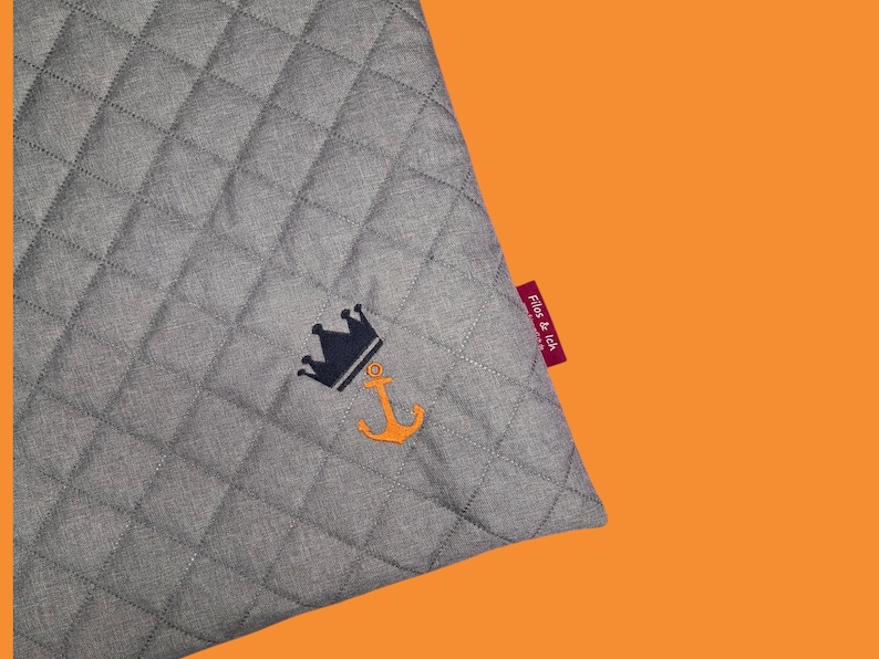 May include: Gray quilted fabric with a navy blue crown and orange anchor embroidery. A red tag with the text "Fios & Ich" is attached to the corner. The background is orange.