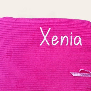 May include: A pink corduroy pillow with the name "Xenia" embroidered in white. The pillow is tied with a white and pink gingham ribbon.