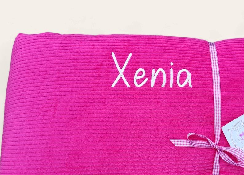 May include: A pink corduroy pillow with the name "Xenia" embroidered in white. The pillow is tied with a white and pink gingham ribbon.