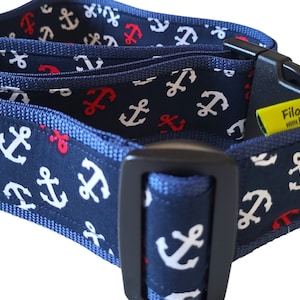 Könnte beinhalten: Ein dunkelblaues Stoffhalsband mit einem sich wiederholenden Muster aus weißen und roten Ankern. Das Halsband verfügt über eine schwarze Kunststoff-Steckschnalle, einen schwarzen Kunststoff-Schieber und ein gelbes Stoffetikett mit der Aufschrift Filos & Ich.