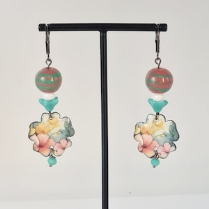 May include: A pair of floral earrings with a pink and green striped bead and a turquoise bead dangle. The earrings are displayed on a black stand.