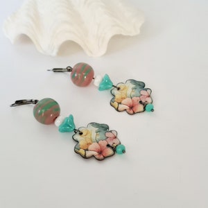 May include: A pair of floral dangle earrings with pink, yellow, and green accents. The earrings feature a floral design with a pink, yellow, and green color scheme. The earrings are made with a silver-toned metal and have a hook closure.