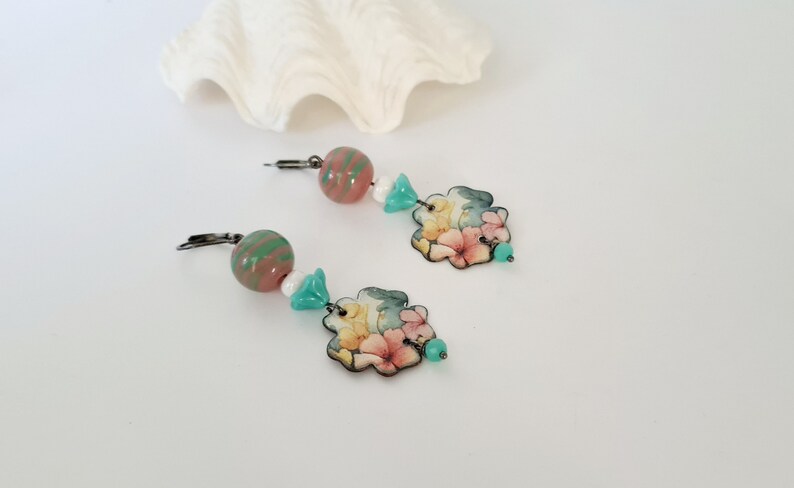 May include: A pair of floral dangle earrings with pink, yellow, and green accents. The earrings feature a floral design with a pink, yellow, and green color scheme. The earrings are made with a silver-toned metal and have a hook closure.