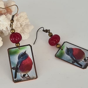 Op de afbeelding: Een paar oorhangers met een rode glazen kraal en een rechthoekige koperen hanger met een kleurrijk beeld van een vogel.