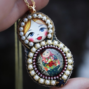 May include: A gold necklace with a pendant featuring two nested Russian nesting dolls. The dolls are painted with colorful faces and traditional clothing. The pendant is surrounded by white pearls.