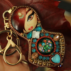 Peut inclure: Un porte-clés doré avec un design coloré et orné de bijoux. Au centre, un portrait peint d'une femme aux cheveux roux et aux yeux bleus. Le porte-clés est orné de perles turquoise, rouges et dorées.