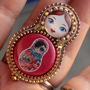 May include: A red and gold beaded brooch with a round, red centre featuring a colourful image of a Russian nesting doll. The doll has a red dress and a black and white patterned skirt.
