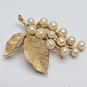 May include: A gold-coloured brooch shaped like a branch with leaves and faux pearls. The brooch features a cluster of cream-coloured pearls and detailed leaf accents, creating a vintage aesthetic. The brooch is set against a white background.