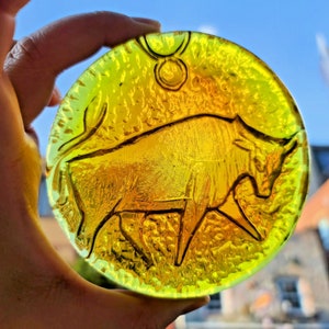 Puede incluir: Adorno redondo de vidrio teñido de amarillo con una detallada representación de un toro, el símbolo del zodiaco Tauro. El vidrio texturizado se sostiene contra un cielo azul brillante, con un edificio al fondo.