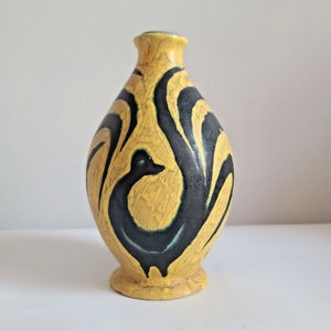 Könnte beinhalten: Eine dekorative Keramikvase mit gelbem Grund und schwarzem, abstraktem Design. Die Vase hat einen abgerundeten Körper, einen schmalen Hals und einen kleinen, kreisförmigen Sockel. Das schwarze Design ähnelt einem Vogel. Die Vase ist etwa 20 cm hoch.