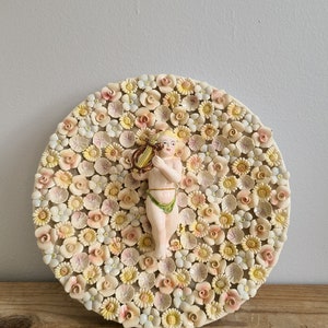 May include: A round ceramic plate decorated with a floral design featuring a variety of pink, white, and yellow flowers. A small ceramic figurine of a cherub playing a lyre is centered on the plate.
