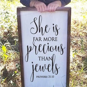 May include: A framed wooden sign with the text "She is far more precious than jewels" in elegant script. The sign also includes the Bible verse "Proverbs 31:10". The frame is a dark brown color, and the background is a light cream color.