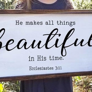 May include: A white wooden sign with brown wood frame, featuring the text "He makes all things beautiful in His time. Ecclesiastes 3:11".
