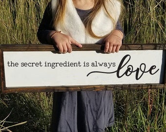 May include: A white wooden sign with brown trim reads "the secret ingredient is always love" in black lettering. The sign is held by a person in front of a grassy background.