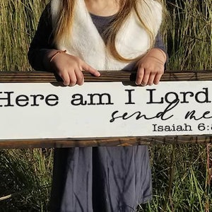 May include: A rectangular wooden sign with the text "Here am I Lord, send me" and the reference "Isaiah 6:8" in black lettering. The sign has a white background and a dark brown wooden frame. The sign is held in front of a background of tall grass.