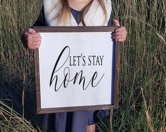 May include: A framed wooden sign with the words "Let's Stay Home" in black script on a white background. The sign is held in front of a background of tall grass.