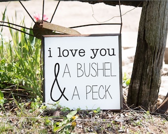 May include: A white wooden sign with the phrase "i love you A BUSHEL & A PECK" in black lettering. The sign is framed in a dark brown border and is hanging outdoors near greenery and a wooden post.