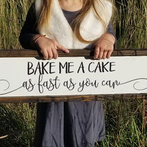 May include: A white wooden sign with brown trim reads "Bake me a cake as fast as you can." The sign is held by a child wearing a white fur vest and a gray dress.