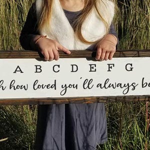 May include: A white wooden sign with a brown frame. The sign says "A B C D E F G oh how loved you'll always be".